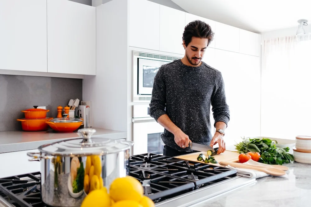 A men are cooking in a modern kitchen, slicing fresh vegetables on a cutting board with bright natural light in the background.