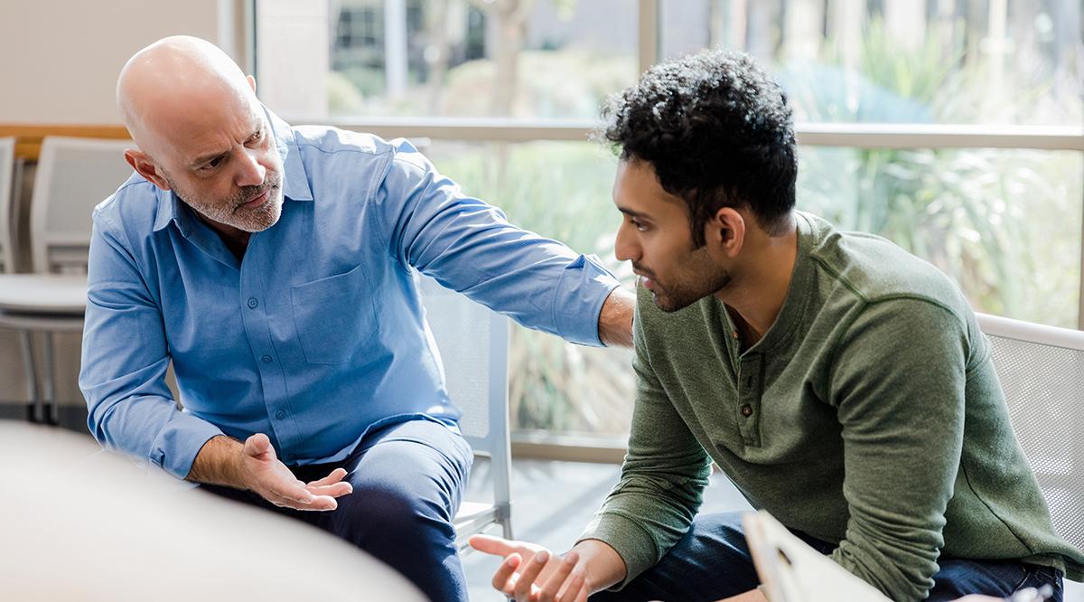 Older man in a blue shirt providing empathetic support to a younger man in a green shirt, emphasizing the importance of men's mental health.