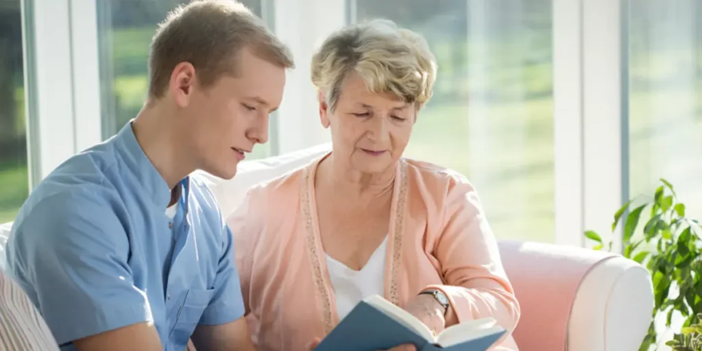 A support worker helping an elderly woman read a book at home, providing care and companionship.