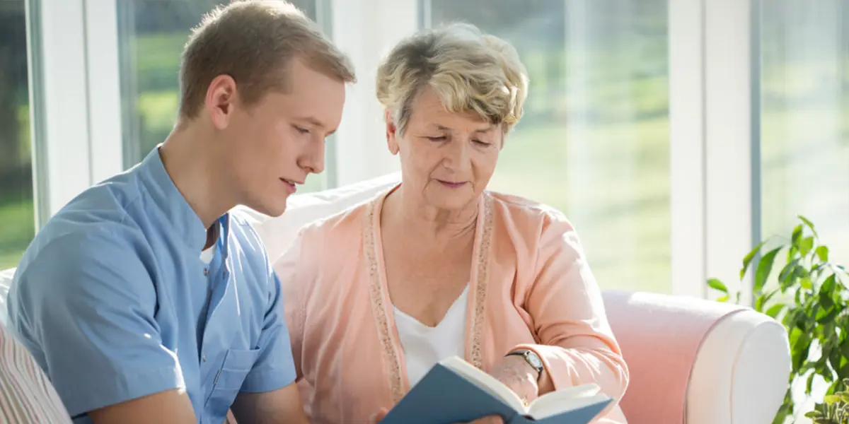 A support worker helping an elderly woman read a book at home, providing care and companionship.
